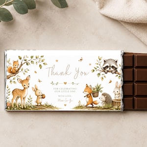 May include: A rectangular chocolate bar wrapped in a white paper label featuring watercolor illustrations of a deer, fox, raccoon, rabbit, hedgehog, and squirrel. The text reads: Thank You FOR CELEBRATING OUR LITTLE ONE WITH LOVE, Mom-to-be.