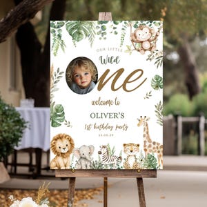 May include: A rectangular birthday party sign featuring watercolour jungle animals including a lion, elephant, zebra, leopard, giraffe, and monkey. The text reads Our Little Wild One, welcome to OLIVER'S 1st birthday party, and 15.05.29.