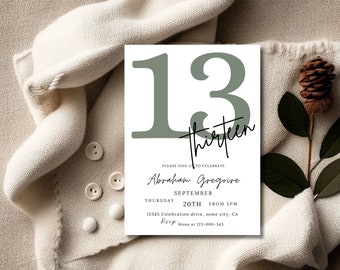 May include: A white invitation card with the number "13" in a muted green color, along with the word "thirteen" in a script font. The card includes event details: name, date, time, and location. The card is placed on a cream-colored fabric.
