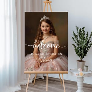 以下が含まれることがあります： イーゼルに立てられた肖像画スタイルのサインで、ピンクのガウンとティアラを着た若い人が特徴です。サインには「welcome to my sweet sixteen」と「Olivia」という名前と「December 12, 202X」の日付が書かれています。