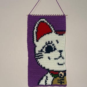 May include: A purple crocheted wall hanging with a white cat face with red ears and a black and gold collar.