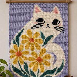 Peut inclure: Une tenture murale en crochet rectangulaire représentant un chat blanc aux oreilles et joues roses, entouré de marguerites jaunes à cœur orange et de feuilles vert foncé, sur fond violet clair, suspendue à une tige en bois.