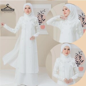May include: A white, long-sleeved dress with floral embroidery and a matching head covering. The dress features a flowing skirt and delicate embellishments, creating an elegant and modest look. The brand logo "AYRA & AYDAN" is visible in the top left corner.