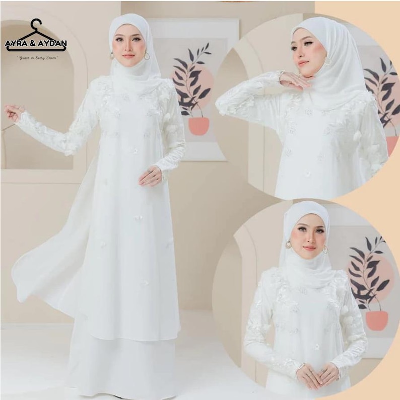 May include: A white, long-sleeved dress with floral embroidery and a matching head covering. The dress features a flowing skirt and delicate embellishments, creating an elegant and modest look. The brand logo "AYRA & AYDAN" is visible in the top left corner.