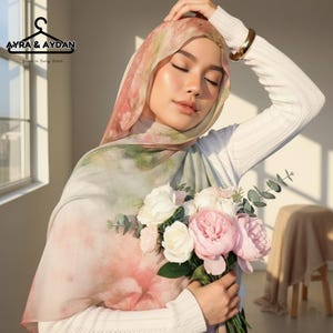 May include: A floral patterned hijab in shades of pink, green, and white. The hijab is draped over the head and shoulders. The image also features a bouquet of white and pink flowers. The brand name "AYRA & AYDAN" is visible in the upper left corner.