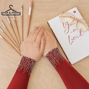 May include: A pair of burgundy sleeves with a delicate white beaded design at the cuffs. The image also features makeup brushes, a book with the words "You are Loved", and eyeglasses. The brand logo "AYRA & AYDAN" is visible.