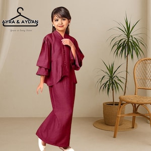 Può includere: Una giovane persona indossa un completo bordeaux a due pezzi. La parte superiore ha un design a maniche a strati e chiusura con bottoni. La gonna è in tinta unita e leggermente svasata. Il marchio "AYRA & AYDAN" è visibile in alto a sinistra.