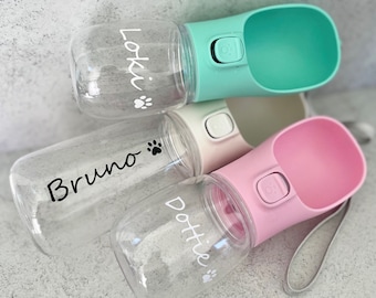 May include: Three clear plastic water bottles with a paw print and a name on each. The bottles have a built-in bowl and a carrying strap. The bottles are in the colors pink, green, and white.