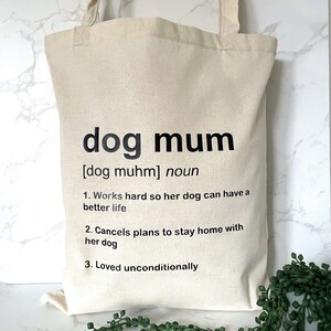 May include: A beige tote bag with black text that reads "dog mum [dog muhm] noun 1. Works hard so her dog can have a better life 2. Cancels plans to stay home with her dog 3. Loved unconditionally"