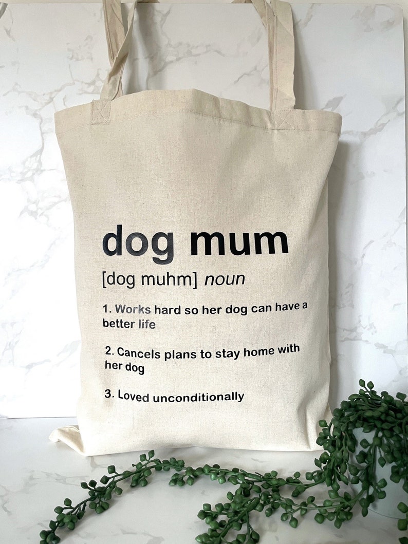 May include: A beige tote bag with black text that reads "dog mum [dog muhm] noun 1. Works hard so her dog can have a better life 2. Cancels plans to stay home with her dog 3. Loved unconditionally"