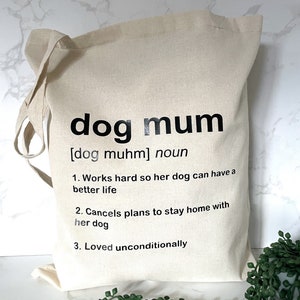 May include: A white canvas tote bag with black text that reads "dog mum [dog muhm] noun 1. Works hard so her dog can have a better life 2. Cancels plans to stay home with her dog 3. Loved unconditionally"