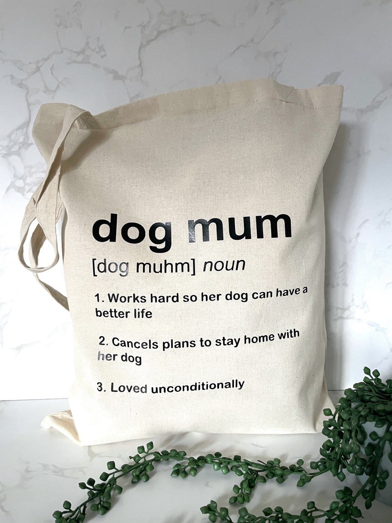 May include: A white canvas tote bag with black text that reads "dog mum [dog muhm] noun 1. Works hard so her dog can have a better life 2. Cancels plans to stay home with her dog 3. Loved unconditionally"