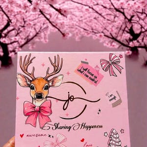 Könnte beinhalten: Eine rosa Box mit einer Hirschillustration mit rosa Schleife und Geweih. Der Text "Sharing Happiness" ist vorhanden, zusammen mit den Sätzen "awesome", "love surrounds me" und "I am enough."