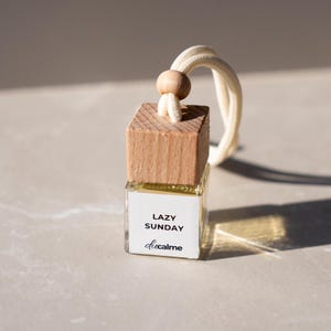 Könnte beinhalten: Kleine, transparente Glasflasche mit Holzdeckel und weißer Kordel. Auf der Flasche steht "LAZY SUNDAY" und "docalme". Der Holzdeckel ist quadratisch und hat eine kleine Holzperle an der Kordel.