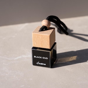 Könnte beinhalten: Ein schwarzer Auto-Lufterfrischer mit einem Holzquader-Aufsatz und einer schwarzen Schnur. Der untere Teil ist ein schwarzes Quadrat mit dem Text "BLACK OUD" und "ducalme" in Weiß. Der Holzquader hat eine hellbraune Farbe.