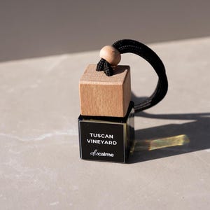 Könnte beinhalten: Aromatische Auto-Lufterfrischer mit einem Holzquader-Aufsatz und einer schwarzen Basis. Die Basis hat ein schwarzes Etikett mit dem Text "Tuscan Vineyard" und dem Markennamen "ducalme" in Weiß. Eine schwarze Kordel und eine Holzperle sind zum Aufhängen befestigt.