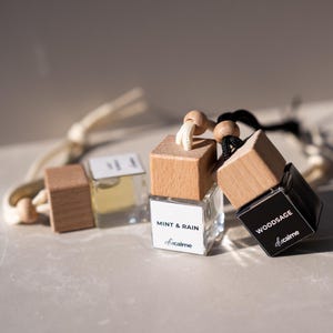 Könnte beinhalten: Drei kleine, quadratische Glasflaschen mit Holzdeckeln und Aufhängeschnüren. Eine Flasche ist schwarz mit dem Wort "WOODSAGE" auf einem weißen Etikett. Eine andere hat ein weißes Etikett mit der Aufschrift "MINT & RAIN". Es sind Lufterfrischer für Autos.