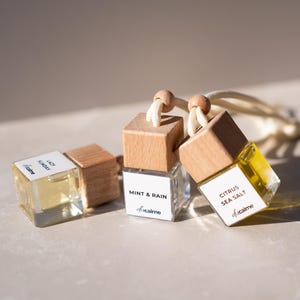 Könnte beinhalten: Drei kleine Glasflaschen mit Holzdeckeln und weißen Etiketten. Auf den Etiketten steht "Lazy Sunday", "Mint & Rain" und "Citrus Sea Salt". Jede Flasche enthält eine gelbe Flüssigkeit und ist an einer weißen Schnur aufgehängt. Der Markenname "dicalme" ist ebenfalls sichtbar.