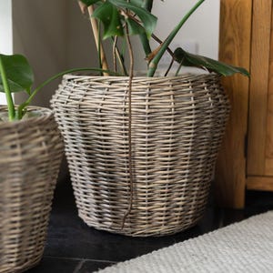 May include: A large, woven wicker basket planter in a natural, light grey colour. The basket has a rounded shape and is filled with green leafy plants. The basket is sitting on a dark surface.