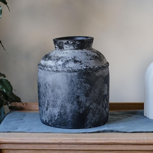 May include: A dark grey decorative vase with a textured, weathered appearance. The vase has a rounded body, a narrow neck, and a slightly flared rim. It sits on a wooden surface.