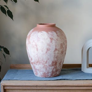 May include: A large, terracotta-coloured vase with a wide body and a narrow neck. The vase has a textured surface with white paint splotches. It sits on a wooden surface covered with a blue cloth. The vase is a decorative home decor item.