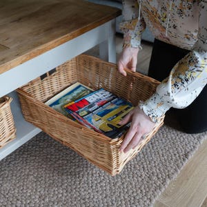 May include: A rectangular wicker basket filled with magazines, positioned under a wooden table. The basket is light brown and has a woven design. The magazines have colourful covers, with yellow and red being the most prominent colours.