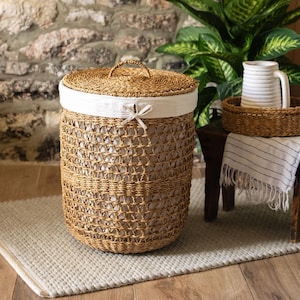 May include: A natural-toned woven laundry basket with a lid and a white fabric liner. The basket has a handle on the lid. The white liner is visible at the top, tied with a bow. The basket sits on a light grey rug.