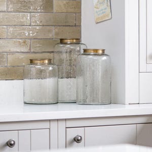 May include: Three clear glass jars with textured sides and gold-coloured lids. The jars are different sizes and sit on a white countertop. The jars are likely for kitchen storage.