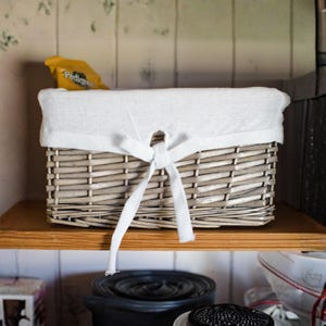 May include: A rectangular wicker basket, light grey in colour, with a white fabric liner and bow. The basket sits on a wooden shelf. A yellow bag of dog food is visible in the background.