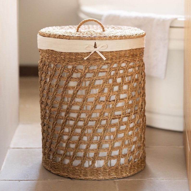 May include: A woven laundry hamper with a lid and a handle. The hamper is made of natural-colored woven material with a lattice design. The lid is also woven and has a white fabric lining. A white bow is tied on the front of the lid.