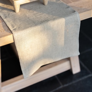 May include: A beige linen table runner on a light-coloured wooden table. The runner has a textured appearance and hangs over the edge of the table. The table is supported by wooden legs, with a dark background.