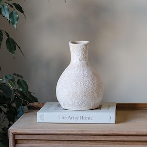 May include: An off-white textured vase with a narrow neck and rounded body. It sits on a book titled "The Art of Home." The vase has a rough, organic texture. The book is on a wooden surface.