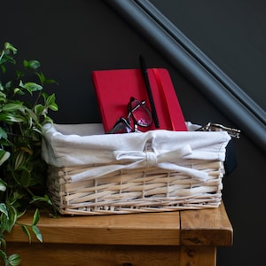 May include: A light-coloured wicker basket with a cream fabric lining and bow. Inside, a red notebook, black pen, and spectacles are visible. The basket is on a wooden surface, with greenery and a dark grey wall behind.