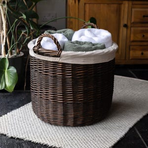 May include: A dark brown wicker laundry basket with a woven handle, filled with rolled white and green towels. The basket sits on a light grey woven rug. The background features plants and a wooden cabinet.