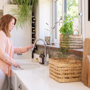 May include: A kitchen interior with a woman at a white sink and chrome tap. Two woven baskets are on a wooden windowsill, one with a plant. A cabinet with wine storage and plants are in the background.