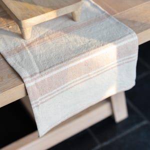 May include: A cream-coloured table runner with horizontal stripes in a muted rose hue. The runner is made of a textured fabric and is draped over a wooden table. A small wooden tray is visible in the background.