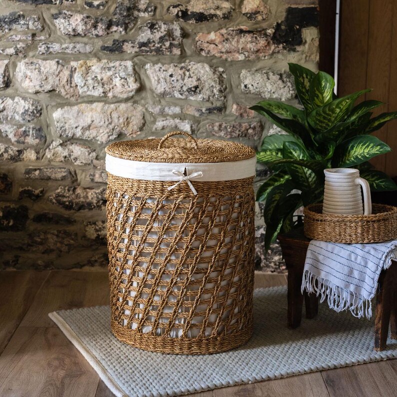 May include: A woven brown laundry hamper with a white fabric liner and lid. The hamper has a diamond pattern and a handle on the lid. A small white bow is tied on the front. The hamper sits on a gray rug.