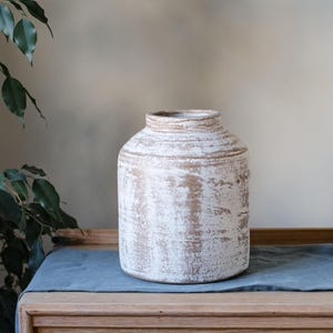May include: A white and gold-toned ceramic vase with a distressed finish. The vase has a wide, rounded body and a slightly narrower neck. The surface has a textured appearance, with visible brushstrokes. The vase is sitting on a wooden surface.