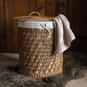 May include: A woven wicker laundry hamper with a lid and a white fabric liner. A beige towel hangs over the side. The hamper is cylindrical and sits on a gray rug. The lid has a handle.