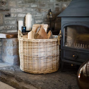 May include: A woven wicker basket, filled with logs, sits on a stone hearth. The basket is light in colour and has two handles. A black fireplace and stone wall are in the background.