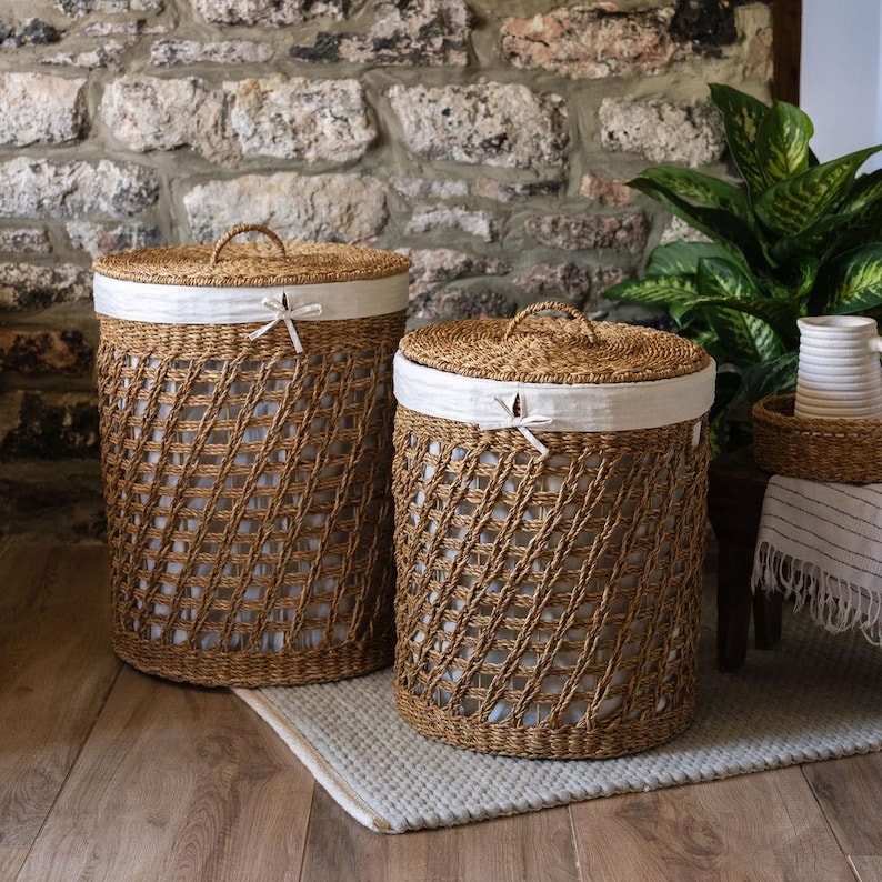 May include: Two woven laundry baskets with lids. The baskets are made of a light brown material with a diamond pattern. Each basket has a white fabric liner and a handle on the lid. The baskets are on a light-colored rug.