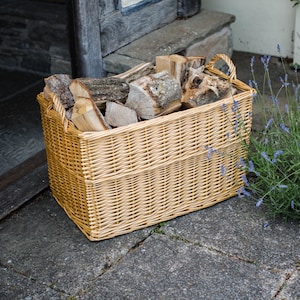 May include: A rectangular wicker basket in a light brown hue, filled with logs of firewood. The basket features two curved handles and is placed on a stone patio near a doorway. The wood is a mix of light and dark brown.