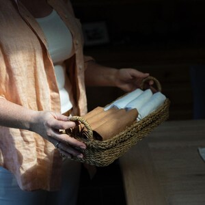 May include: A rectangular wicker basket containing rolled white and brown linens. The basket is held by a person wearing a peach-coloured shirt. The linens are neatly folded and ready for use.