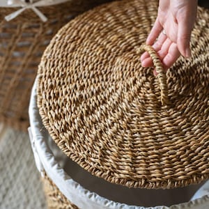 May include: A round woven basket with a lid. The lid is being lifted by a hand. The basket is made of natural-colored woven material. The interior of the basket is lined with a white fabric.