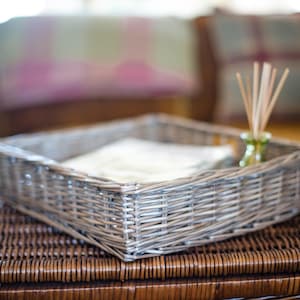 May include: A square, grey wicker tray with a white cloth inside, resting on a brown woven surface. A small green glass bottle with reed diffuser sticks sits to the right of the tray. Decorative cushions are in the background.