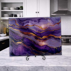 May include: A rectangular glass cutting board featuring an abstract alcohol ink design in shades of deep purple, lavender, and violet, accented with flowing metallic gold veins and glittery gold dust, displayed on a black metal wire stand.