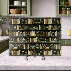May include: A decorative glass panel featuring a bookshelf design with rows of books and floral accents. The panel is supported by a black metal stand and is displayed on a marble countertop in a kitchen setting.