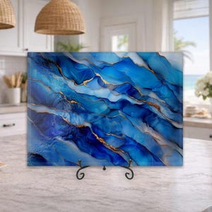 May include: A rectangular glass cutting board featuring a vibrant abstract design of flowing deep blue, light blue, and white waves accented with thin, metallic gold lines. The board is supported by a black metal stand on a kitchen countertop.