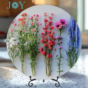 May include: A round decorative plate featuring an arrangement of colorful flowers. The flowers include red, pink, purple, and blue blooms, set against a textured white background. The plate is supported by a black metal stand, ideal for home decor.