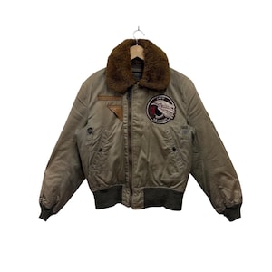 Può includere: Giacca bomber verde oliva con colletto in finta pelliccia marrone. Presenta una toppa con un disegno di aquila e la scritta "343rd Air Apache". La giacca ha orlo e polsini a coste e chiusura con cerniera.